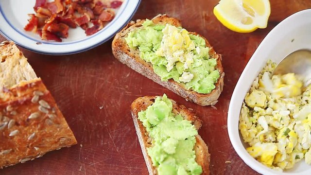 Spooning scrambled eggs onto avocado toast and adding crumbled bacon