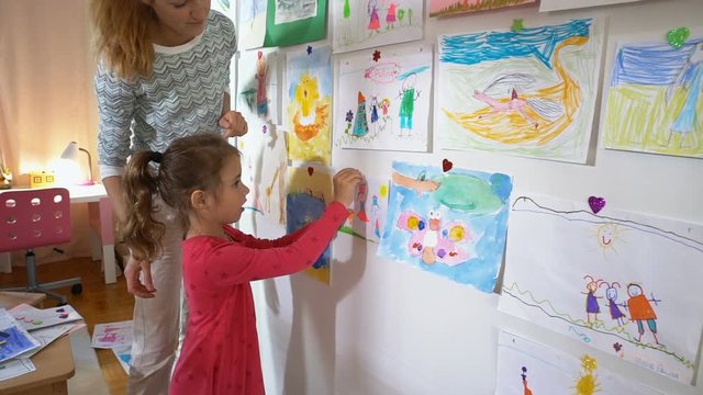 Little cute girl and her mother hang children's drawings on the wall in the nursery. Slow motion.
