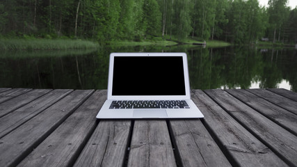 Working with a laptop on a pier, with a forest in the background. This is how we work during the summer in Finland. We use the best technology surrounded by the purest nature.