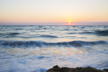 Vista del tramonto dalla spiaggia. Ilmare è calmo e le onde sono poche che si infrangono sulla battigia. Il sole all'orizzonte dona le tinte calde al paesaggio.