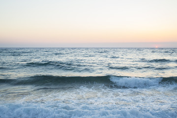 Vista dell'alba dalla spiaggia. Ilmare è calmo e le onde sono poche che si infrangono sulla battigia. Il sole all'orizzonte dona le tinte calde al paesaggio. - obrazy, fototapety, plakaty