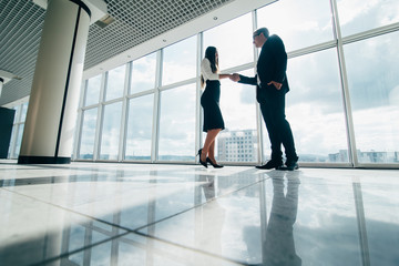 Two smiling young businesspeople shaking hands together in an office building hallway in front of windows overlooking the city
