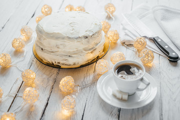 Celebration homemade biscuit cake with white cream on a white wooden table. Twinkling lights around the cake. White cup with black coffee and lump sugar on a saucer. Kitchen knife at the background.