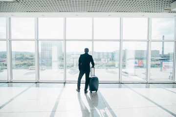 Traveler businessman in airport lounge waiting for the flight and standing with trolley luggage.