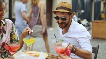 Friends party concept - group of young friends sitting in cafe on the street, portrait of young bearded man with moustache drinking tasty alcohol cocktails in a straw hat, slow motion shot