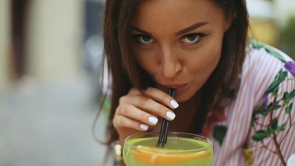 Beautiful girl with hair shaking by wind is drinking a tequila cocktail in the city cafe Bar. Young woman looking at camera, slow motion shot