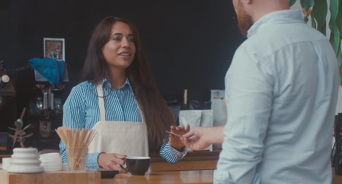 Attractive Caucasian female employee serving a coffee and taking credit card payment from customer at modern coffee shop. 4K UHD 60 FPS