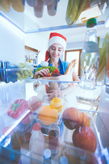 Portrait of female in santa hat standing near open fridge full of healthy food, vegetables and fruits