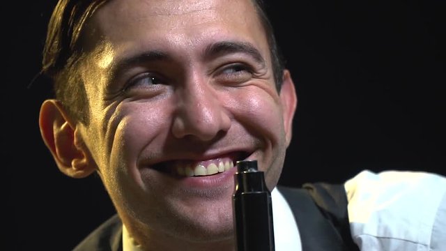 A cheerful man in a suit is crouching holding a gun barrel near the face