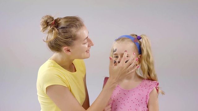A little girl is playing with her mom, a woman is applying paint on her face in order to draw a pet's face