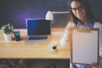Young woman standing near desk with laptop holding folder and cup of coffee. Workplace. Business...