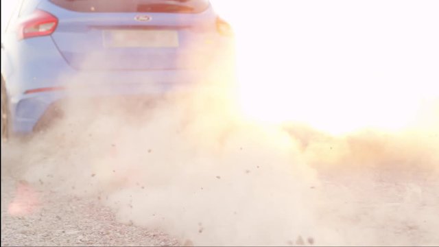 SLOW MOTION CLOSE UP: Sports car starts & quickly drives away from a dusty macadam dirt road raising sand, rocks and stones at dusk. Sportive auto performing burnout at beautiful golden summer sunset