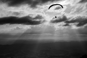 Some para-gliders and hang-gliders flying over a valley, with distant mountains and hills and sun rays coming out through the clouds