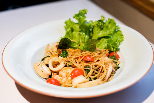 The spaghetti spicy seafood (squid, shrimp) with basil, tomato, black pepper, lettuce and paprika on white dish set on table for food background or texture - homemade food concept.