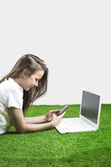 Beautiful smiling happy girl using notebook and mobile smart phone while sitting on the grass. Gray background