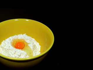 Cracked egg on raw flour in yellow ceramic mixing bowl, black background with free space for text and design 