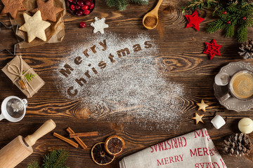Wording Merry Christmas written on dark wooden background, copy space. Christmas concept. Cookies, candle, cinnamon, cup of coffee, marshmallows, fir-tree. View from above.