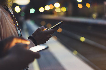 Enjoying travel. Young woman waiting on station platform on background light electric moving train using smart phone in night. Tourist texting message and plan stop railway, railroad transport, bokeh
