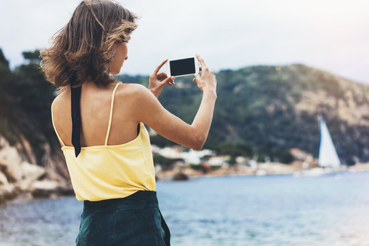 Hipster girl photograph on smart phone gadget in sand coastline, mock up of blank screen. Traveler using in female hand mobile on background seascape horizon. Tourist and blue sun ocean, lifestyle