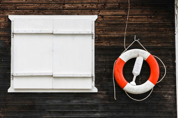Red and white lifebuoy lifebelt with ropes hanging from a dark brown painted wooden wall with a large white window with empty negative copy space on the left of the frame.