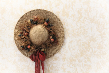 Traditionally decorated vintage handmade lady straw hat with floral bouquet, red ribbon and fruits hanging on a wall decorated with countryside wallpaper.