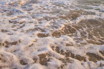Dettaglio della schiuma che creano le onde quando sbattono sul bagnasciuga della spiaggia. L'acqua è trasparente e chiara e attraverso si vede la sabbia del fondale.