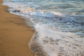 Dettaglio delle onde che si infrangono sulla spiaggia e lasciano la schiuma. La sabbia è dorata e il mare azzurro e agitato.