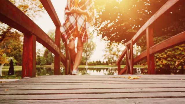 Woman Walking crossing Wooden Bridge in the Autumn Park. 4K. Unrecognizable young woman wearing denim sneakers shoes enjoying calm evening outdoors. Close up on boots. 
