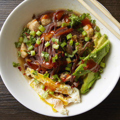 White bowl of chicken and veggie Pho with egg, avocado and siracha