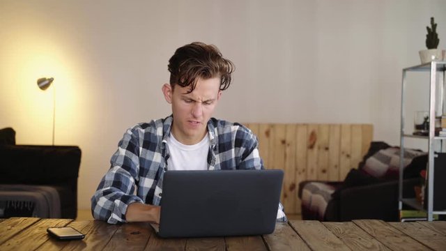 Attractive male millennial hipster in shirt leads an intense conversation with someone through internet online chat, gets annoyed and angry, slams fingers on keyboard