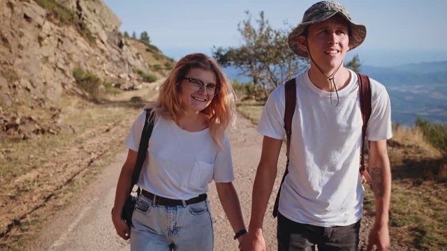 Frontal view on beautiful couple wearing white tshirts, adventure clothing, holding hands and walking on mountain road, enjoying outdoor sport activity together