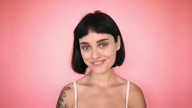 Cute sexy pretty woman in tank top and tattooed arms smiles and smirks flirting to camera, with bright blue eyes and short thick brown hair, on bright coloutful optimistic pink background