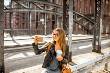 Fototapeta na wymiar Lifestyle portrait of a stylish businesswoman in leather jacket sitting with smartphone and coffeecup on the old iron bridge