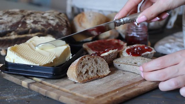 On a brown wooden table lies a breadboard. The slices of bread were on the board. The girl uses a table-knife with a lube-oil knife to spread butter on the bread.