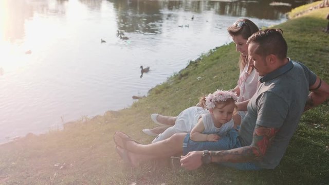 Young family with Baby Girl Enjoying Sunny evening in the Park. Slow Motion 120 fps, 4K. Parents relaxing with their toddler daughter near lake. Happy childhood and Parenthood. Love and Happiness 