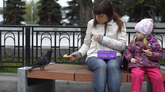 A young family, mother and daughter are eating sandwiches and feeding pigeons sitting on a bench in a public park on a cold, cloudy day.