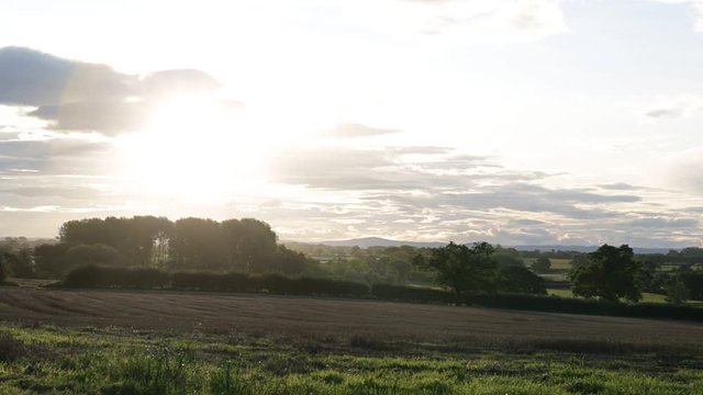 Rural autumnal landscape with brown mowed wheat field and sun setting over horizon, Tarvin, Cheshire, England, UK. Overcast sky with mountains and trees in background. Sunset