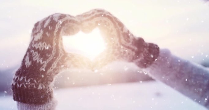 Hands in winter gloves make the shape of heart sign with snow-covered mountains as a background. Slow Motion, 4K DCi. Love and romance on the foreground. Alpine village in the distance.  Love Winter