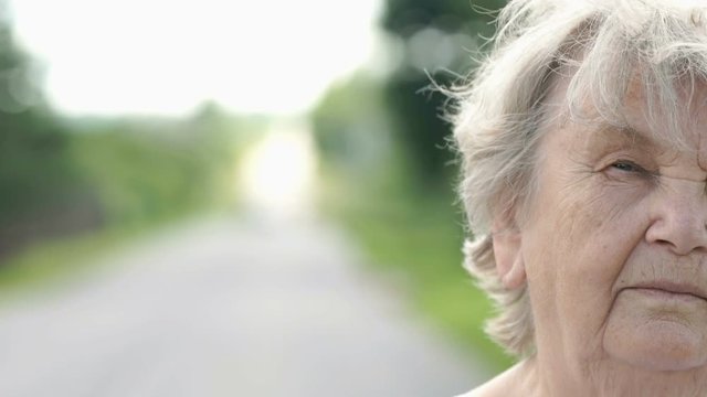 Half face of serious mature elderly woman with gray hair in the background of road in summer. Slow Motion