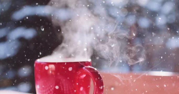 Steaming Cup of Hot Coffee or Tea standing on the Outdoor Table in Snowy Winter Morning. 4K DCi SLOW MOTION 120 fps. Cozy Festive Red Mug with a Warm Drink in Winter Garden. Christmas Morning Concept
