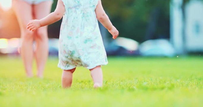 Little baby learns to walk. First Steps. Close Up. SLOW MOTION 120 fps, 4K. Toddler girl running to mother on a green grass, sunny morning or evening. Happy childhood concept. Lens Flare.