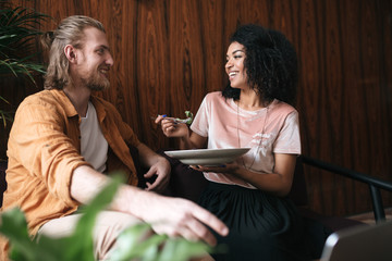 Beautiful young couple sitting and happily talking in restaurant. Pretty African American lady sitting in cafe with plate of salad in hand. Friends having lunch in restaurant