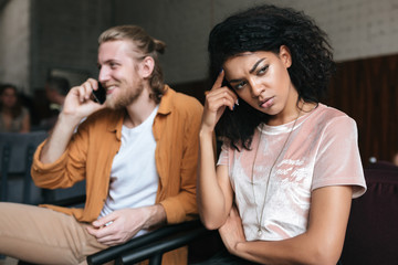 Portrait of young man and girl sitting in restaurant. Angry African American girl with dark curly hair sadly looking aside while boy with blond hair happily talking on his cellphone at cafe
