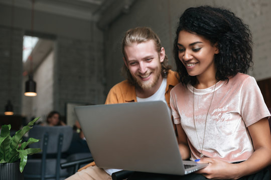 Portrait of young man and girl sitting in restaurant and working on laptop. Nice African American girl sitting with friend and holding laptop in hands at cafe. Boy and girl happily looking at laptop