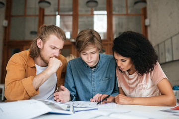 Portrait of young thoughtful people working in office. Two boys with blond hair and girl with dark curly hair sitting and studying together in classroom. Tired students working on new project