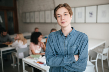 Portrait of young man with blond hair looking in camera in office. Cool boy in denim shirt standing with clasped hands in classroom with students on background