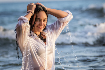 Chica joven con camisa blanca bañándose en el mar al atardecer y jugando con su pelo largo moreno.