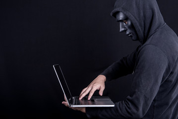 Anonymous male hacker using black mask to cover his face carrying laptop computer. Internet security and cyber attack concepts.