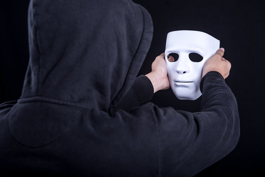 Mystery man holding and looking at white mask. Anonymous social masking or halloween concept.