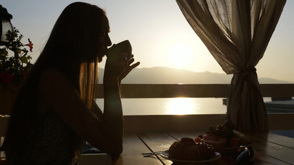 Silhouette of young woman sits in a cafe with panoramic views of sea bay.